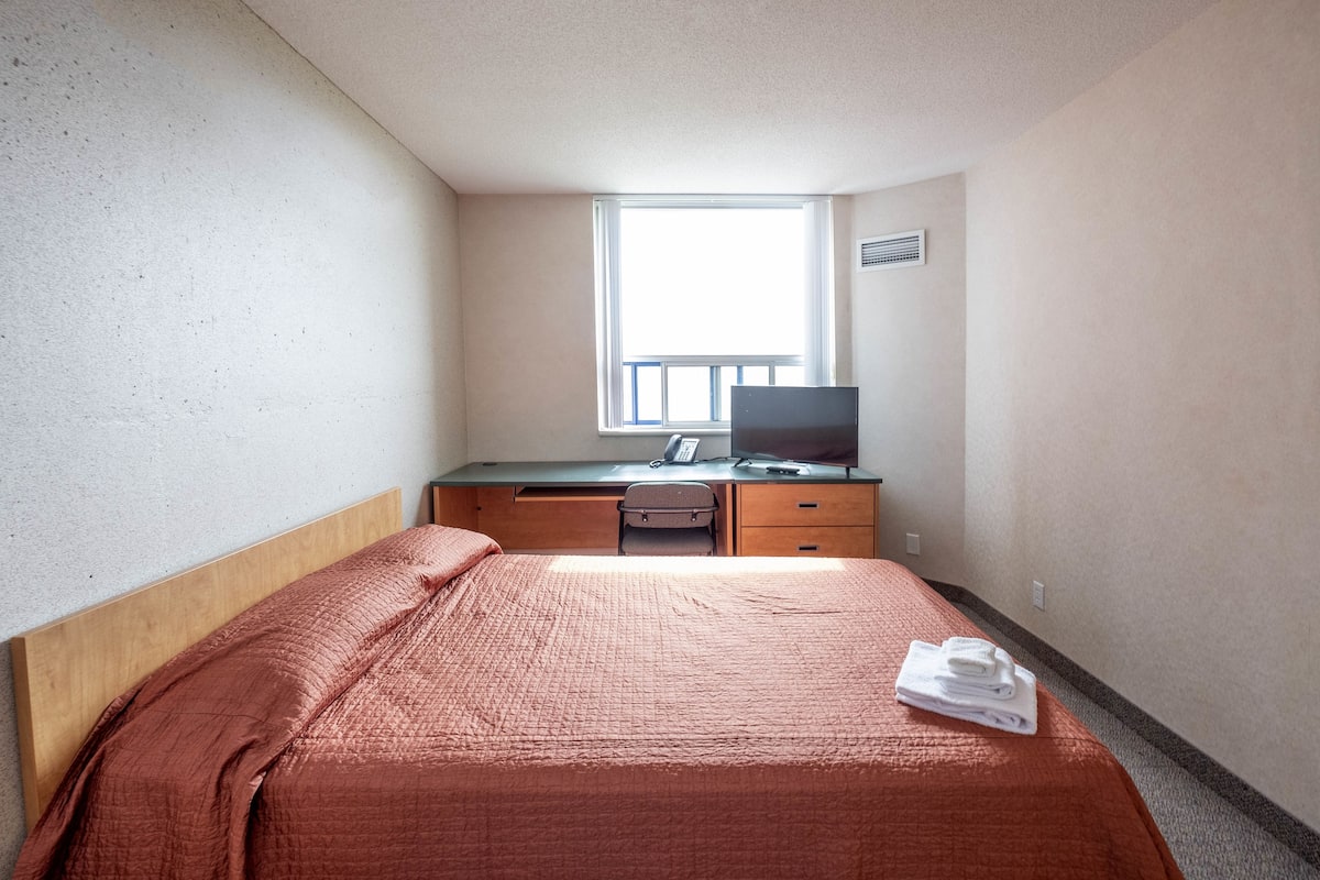 Une chambre d’hôtel avec un lit, un bureau avec un ordinateur, une chaise et une fenêtre.