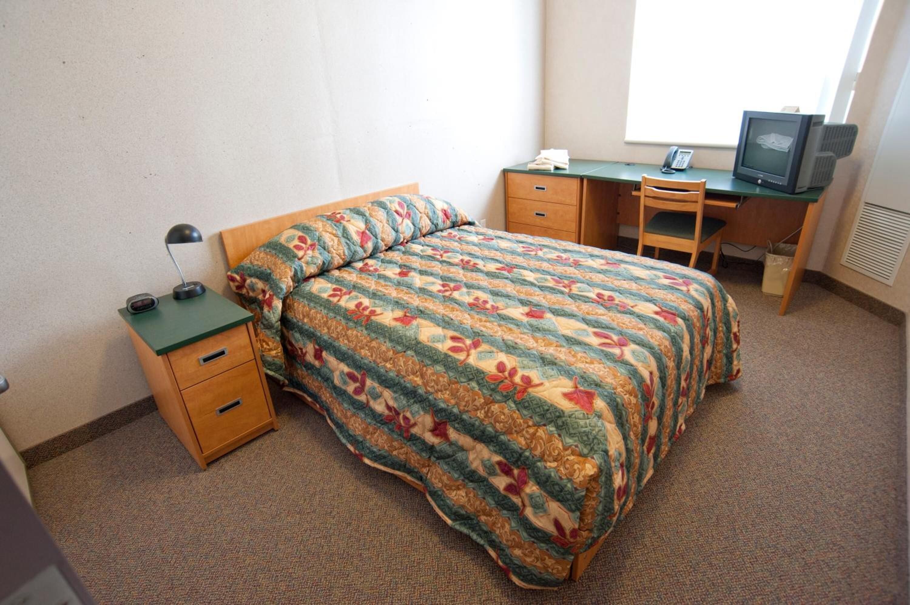 A bedroom with a bed, a desk, a chair, a telephone, and a television.