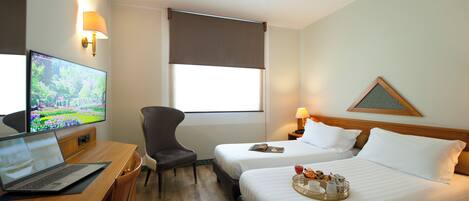 A hotel room with a desk, laptop, TV, two beds, a chair, and a window with a view.