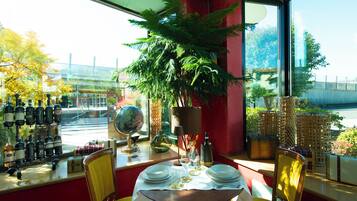 Interior de un restaurante con una mesa para dos, una planta grande en maceta y vista a la calle.
