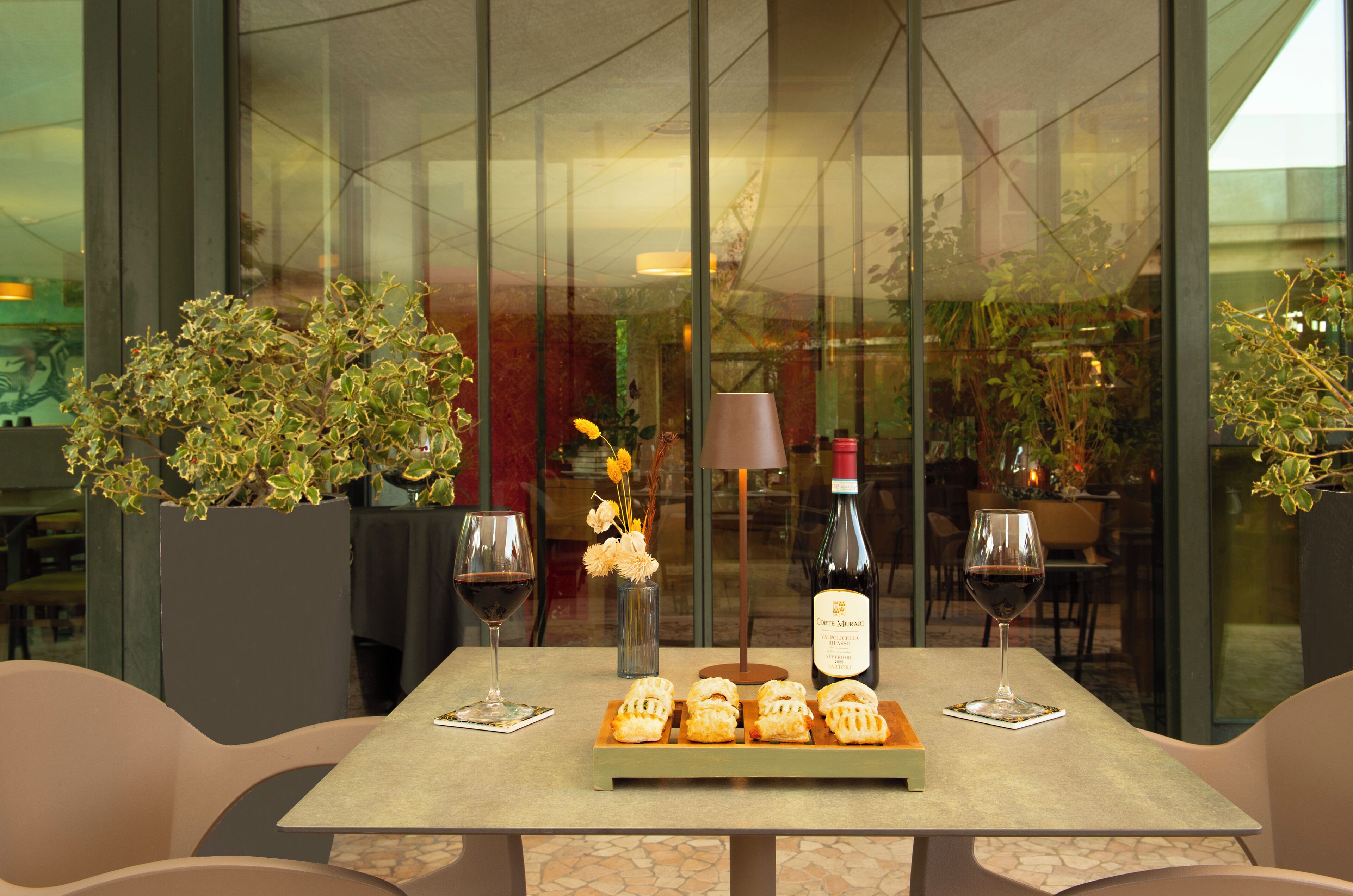 A table with pastries, wine glasses, and a bottle in a modern interior with glass walls and potted plants.