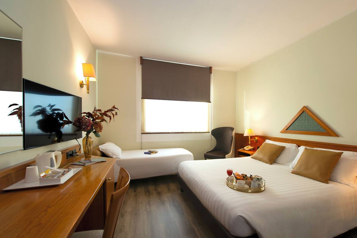 A hotel room with a bed, a desk, a chair, a bedside table with a lamp, a telephone, and a wall-mounted TV.