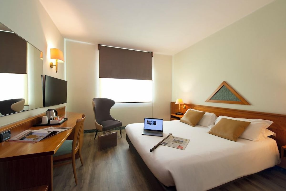 A hotel room with a bed, a desk, a chair, a TV, and a window with a blind.
