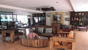 A spacious living area with a large sofa, coffee table, and wooden chairs. There is a TV and a fireplace in the background. - Aiyaree Place Hotel (Pattaya)