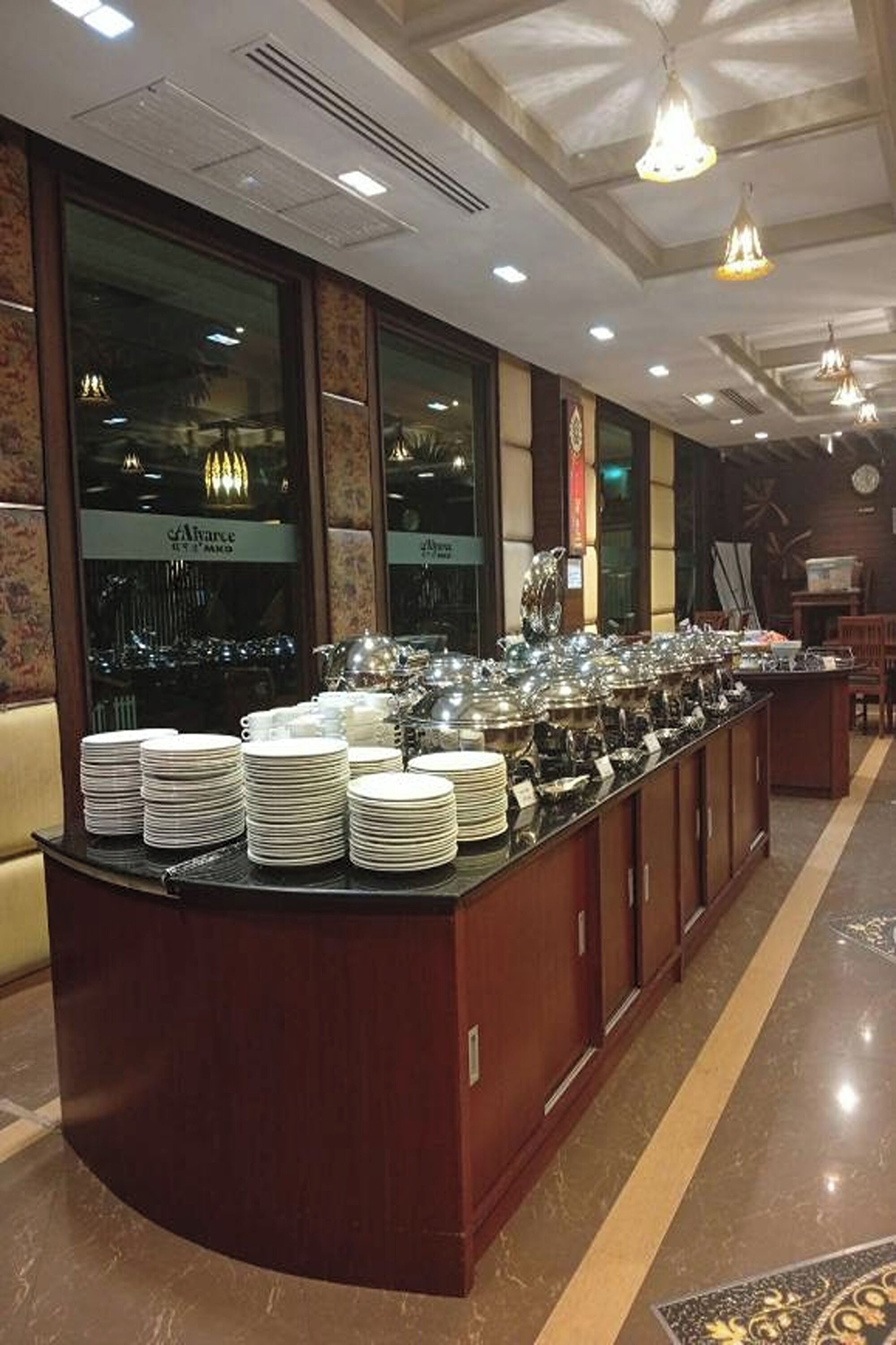 Disposición de un bufé con varias estaciones de servicio, una barra con platos apilados y un interior bien iluminado con luces de techo y ventanas amplias.