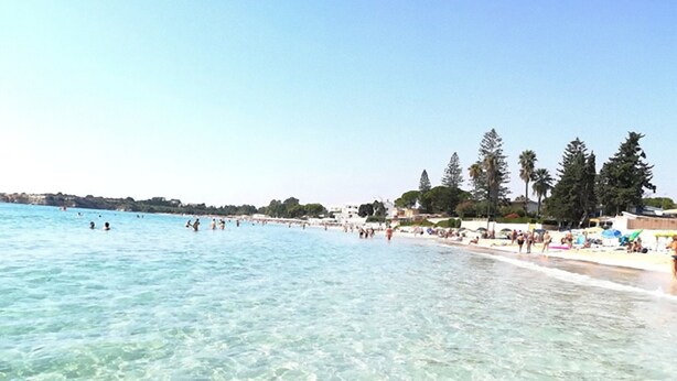 Una playa con agua turquesa cristalina, gente nadando y tomando el sol, y palmeras a lo largo de la orilla.