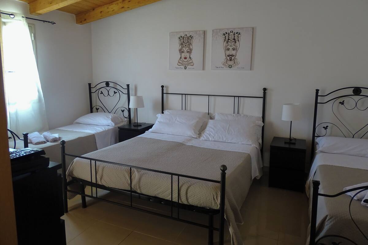 A bedroom with two single beds and a larger bed, all with black metal frames. There are two bedside tables with lamps, and the room has white walls and a wooden ceiling.