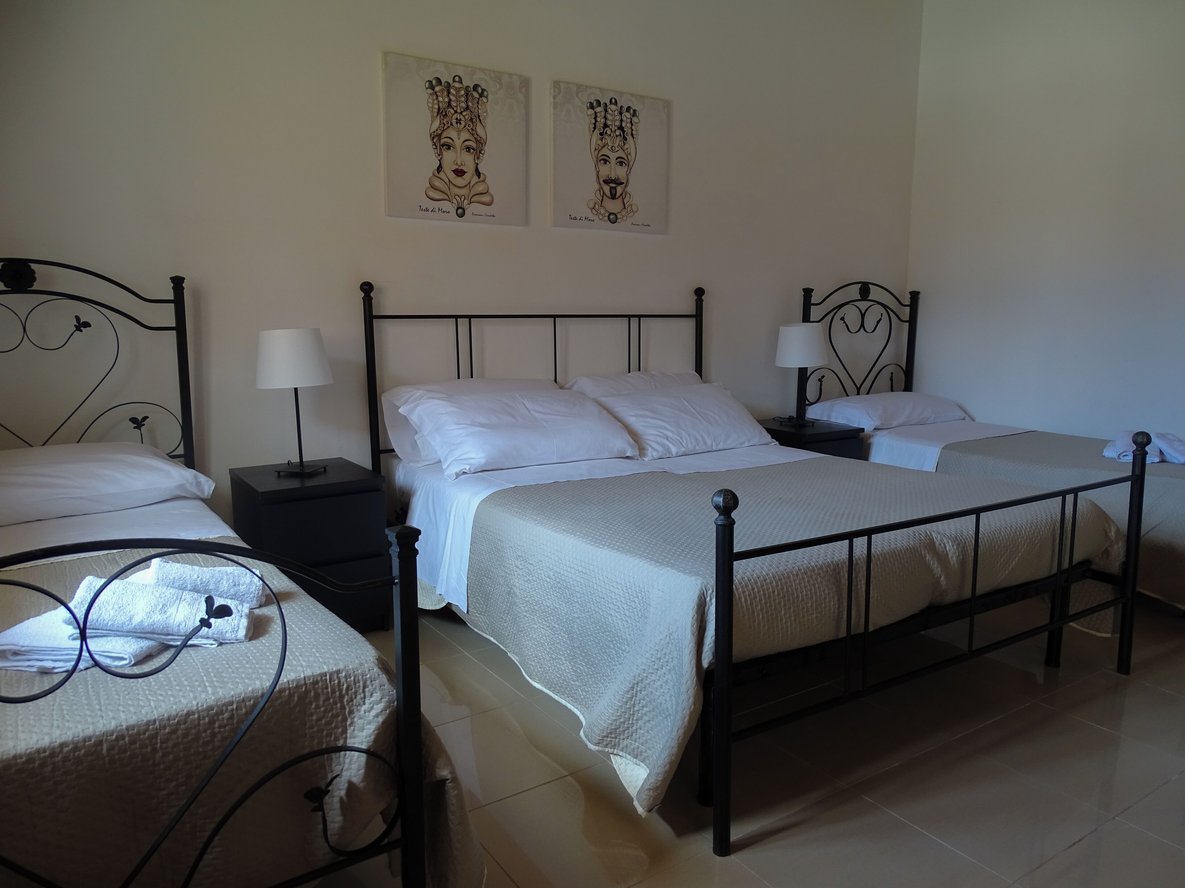 Een hotelkamer met drie eenpersoonsbedden, elk met een zwart metalen frame en witte beddengoed. Er zijn twee nachtkastjes met lampen, en de muren zijn versierd met ingelijste kunstwerken.