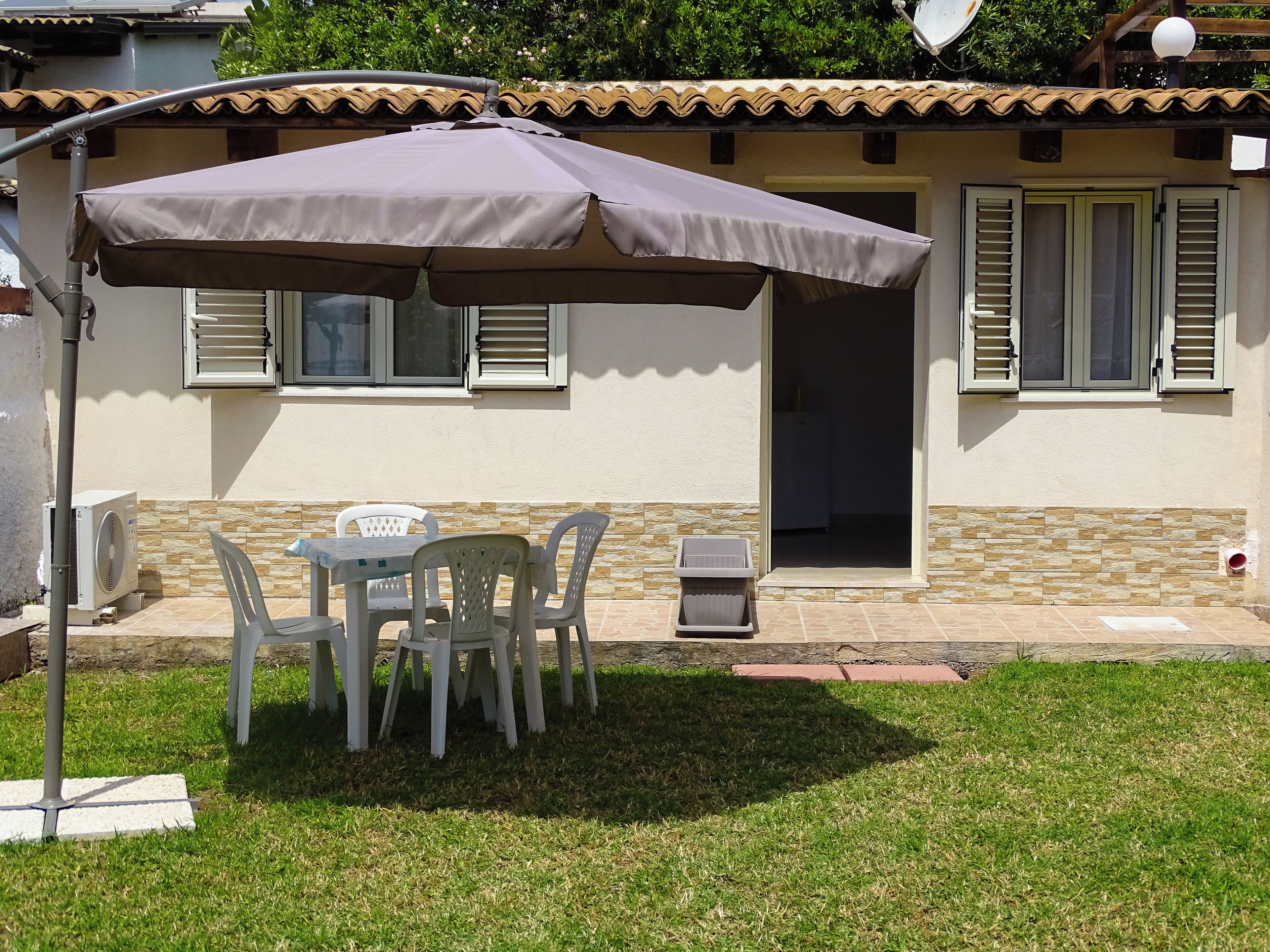 A patio with a table and chairs under a large umbrella, a small trash bin, and a building with a tiled roof and shutters.