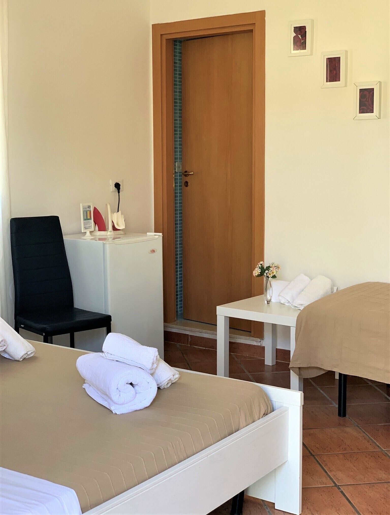 Une chambre d’hôtel avec deux lits, un bureau, une chaise, une petite table ornée d’un vase de fleurs et une porte avec une bande bleue.