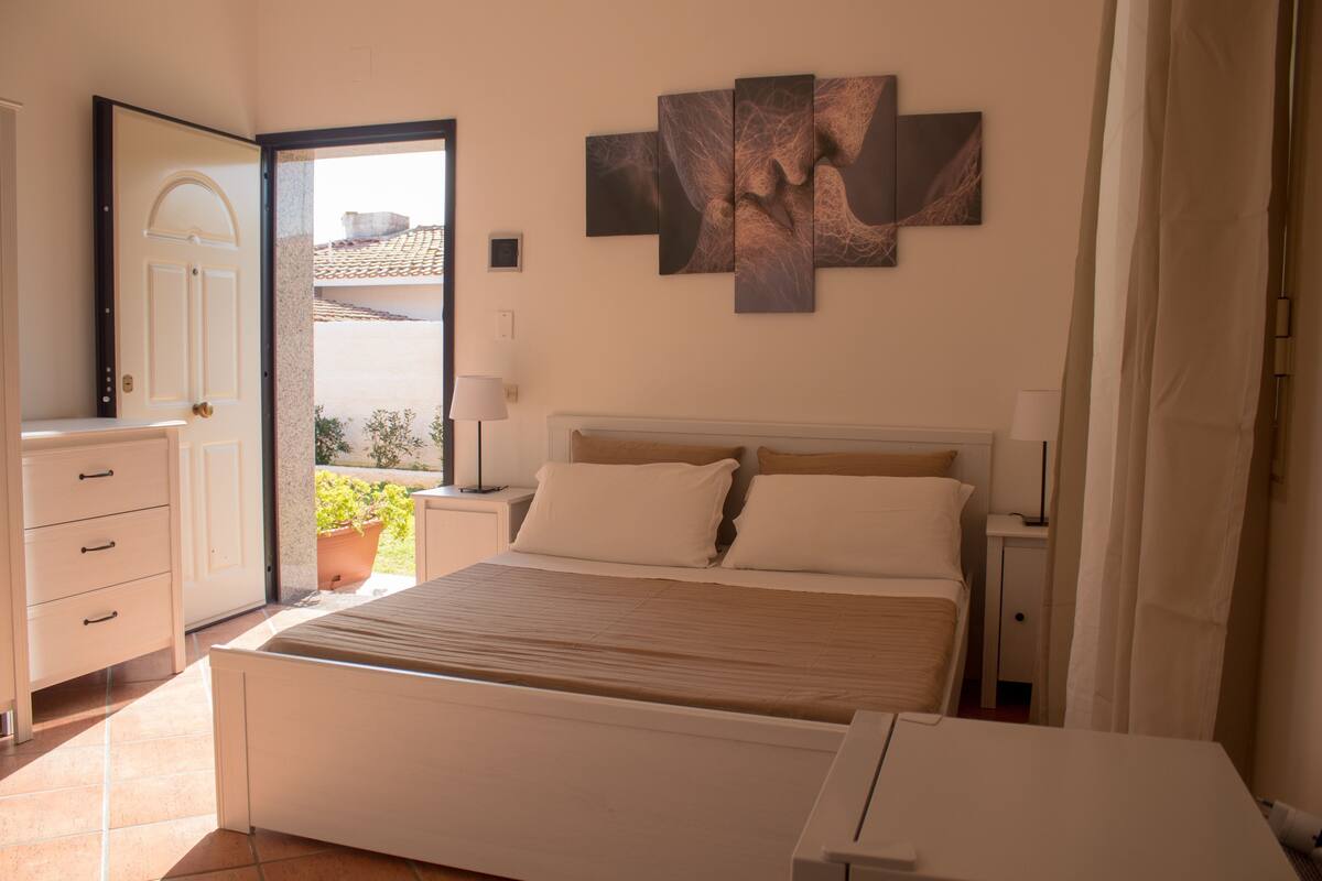 A bedroom with a bed, a nightstand, a lamp, a dresser, and a large window with a view of the outside.