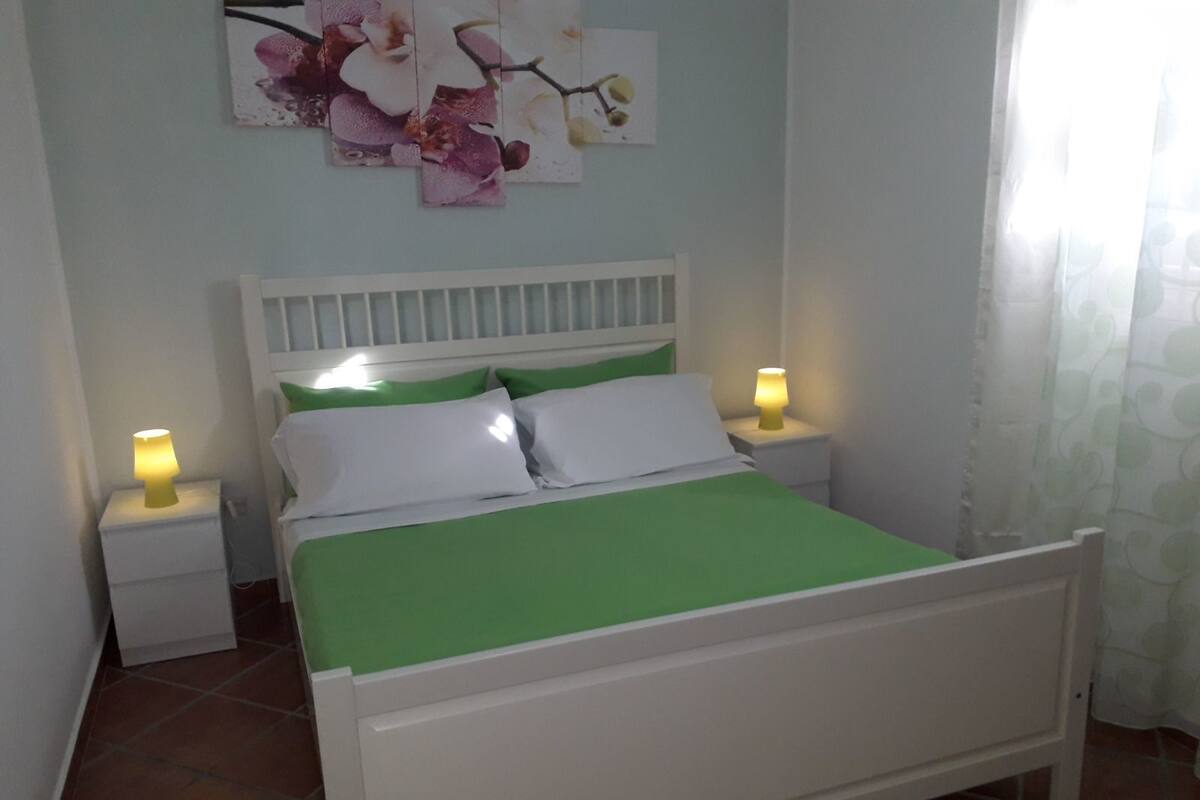 A bed with white headboard and green bedding, two bedside tables with lamps, and a floral artwork on the wall.