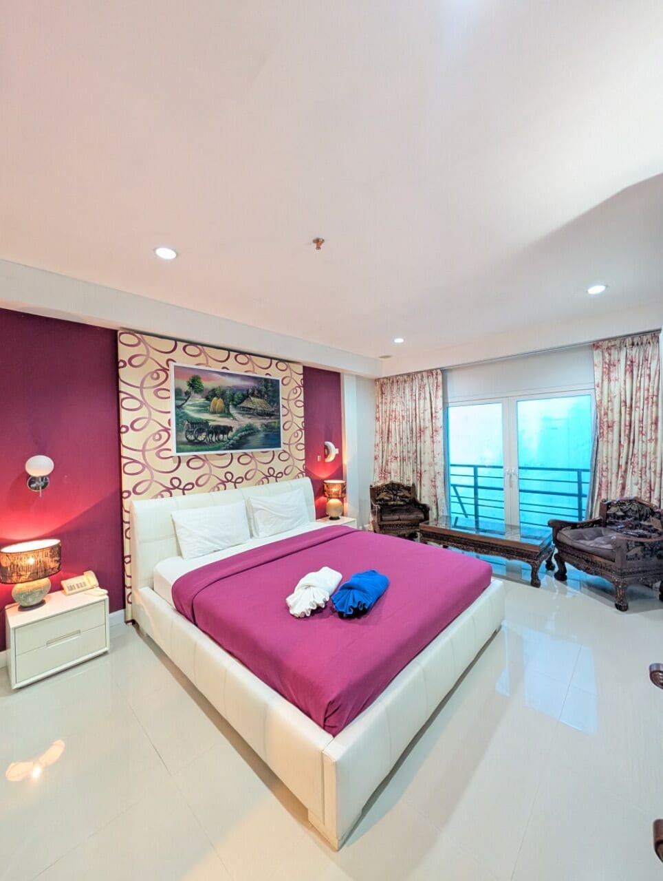 A hotel room with a large bed, a nightstand, a lamp, a chair, and a view of the sea.