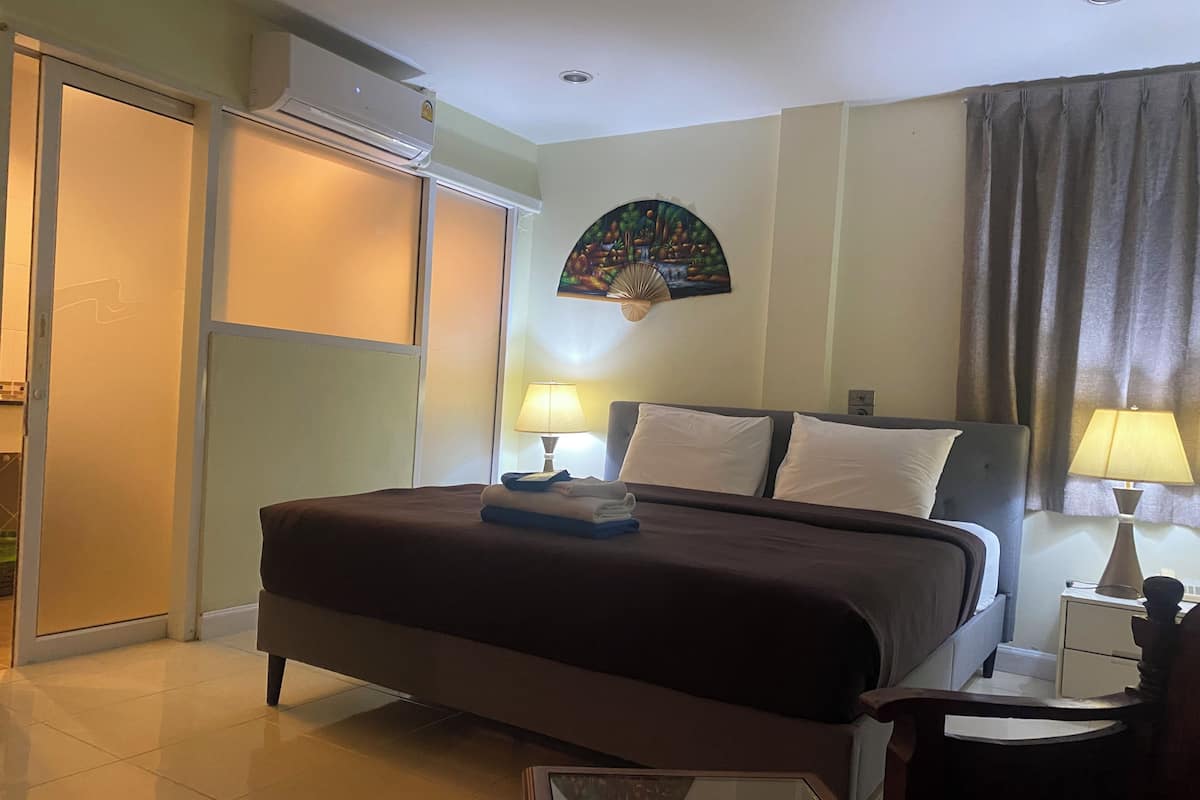 A hotel room with a bed, two bedside lamps, a small table, a chair, a wall-mounted air conditioner, and a decorative wall piece.