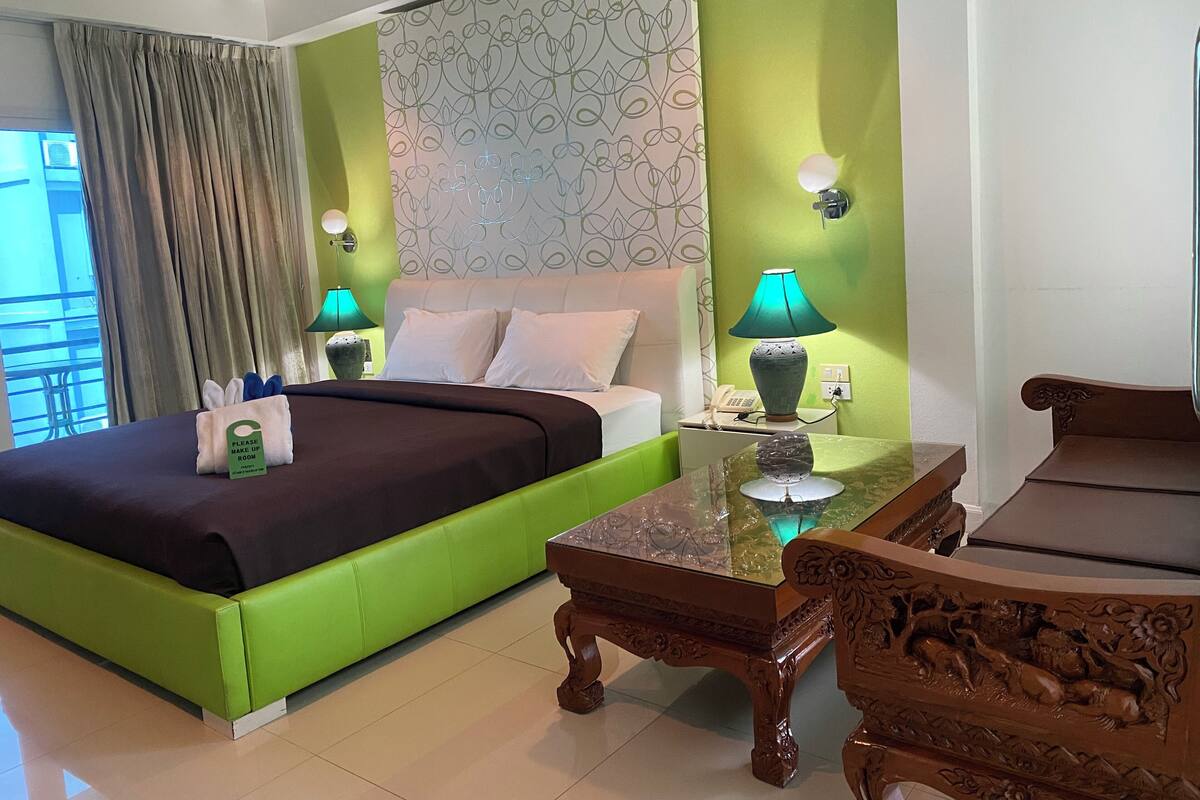 A hotel room with a green bed, a wooden table with carvings, a green lamp, and a patterned wall.
