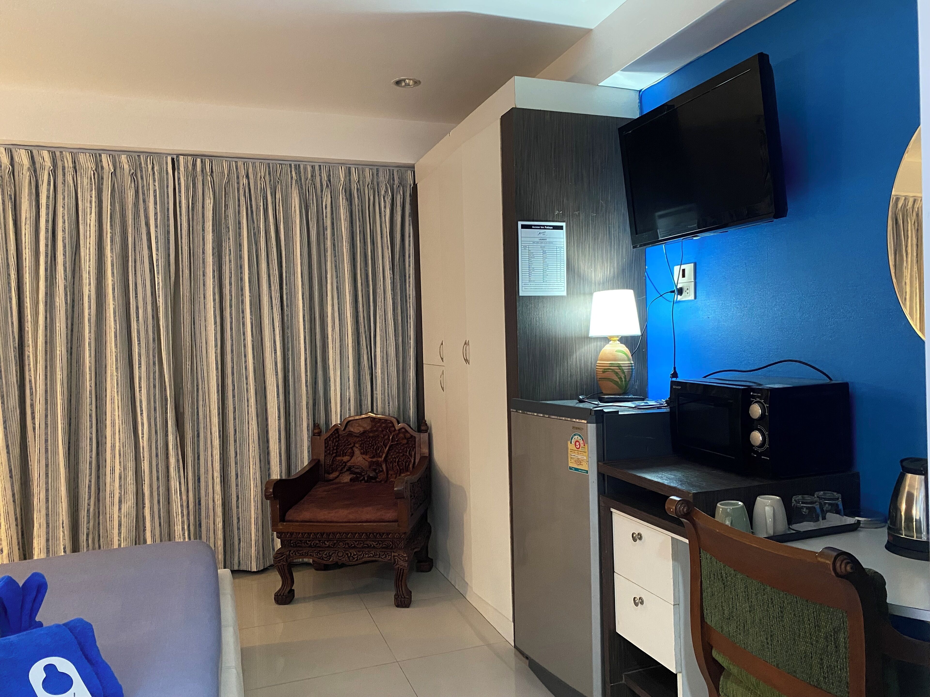 A hotel room with a wooden chair, a desk with a lamp, a flat-screen TV, and a minibar.