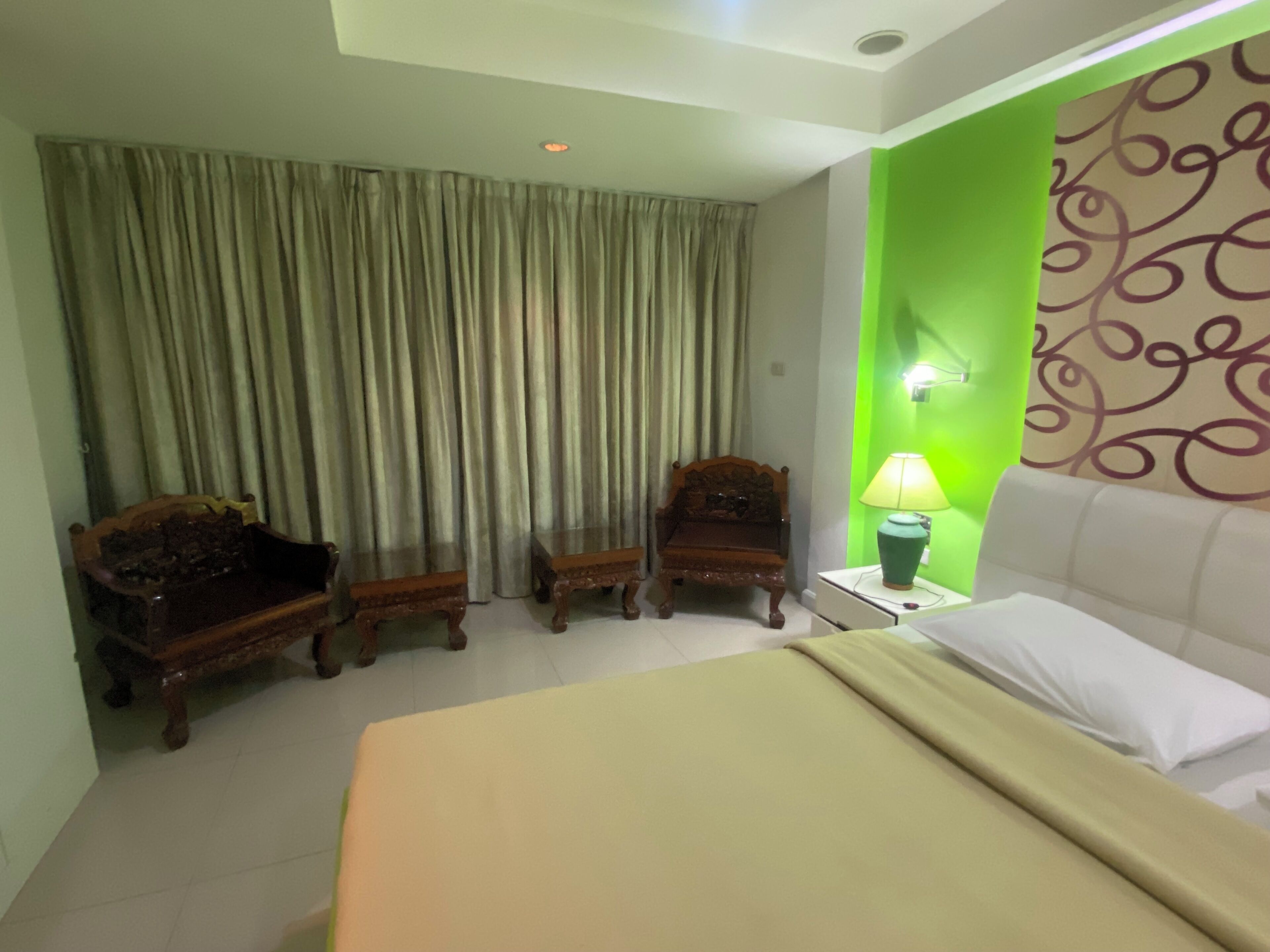 Una habitación de hotel con una cama, dos sillas de madera, una mesita de noche con lámpara y una pared verde decorativa.