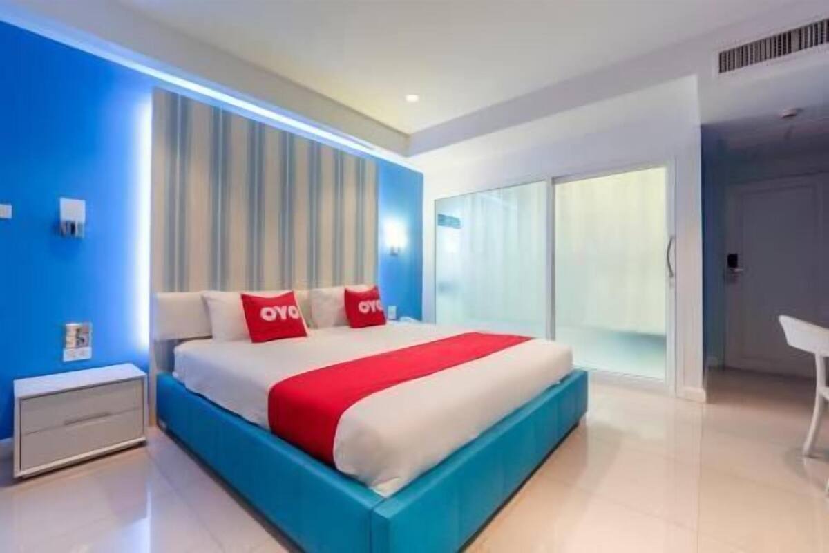 A modern bedroom with a large bed, a nightstand, a desk, and a sliding glass door to a balcony.