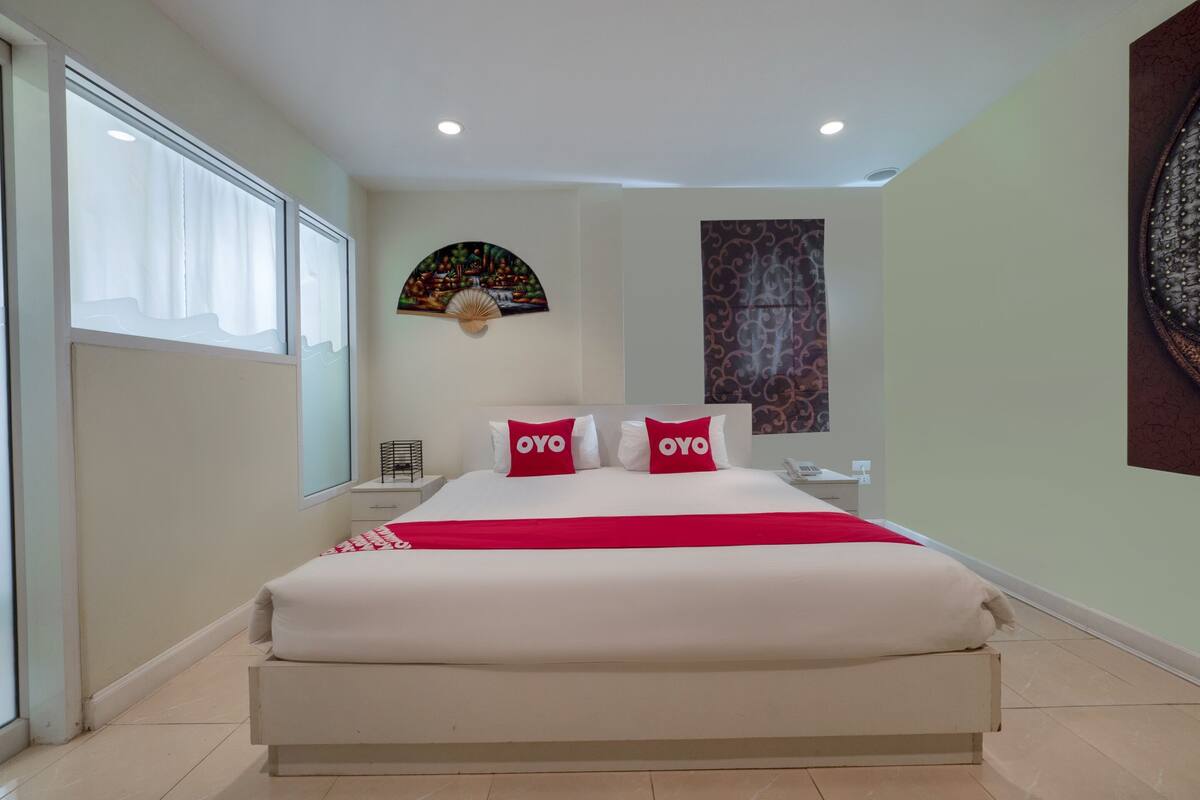 A hotel room with a large bed, two pillows with 'OYO' branding, a nightstand, a wall-mounted fan, and a large window with curtains.