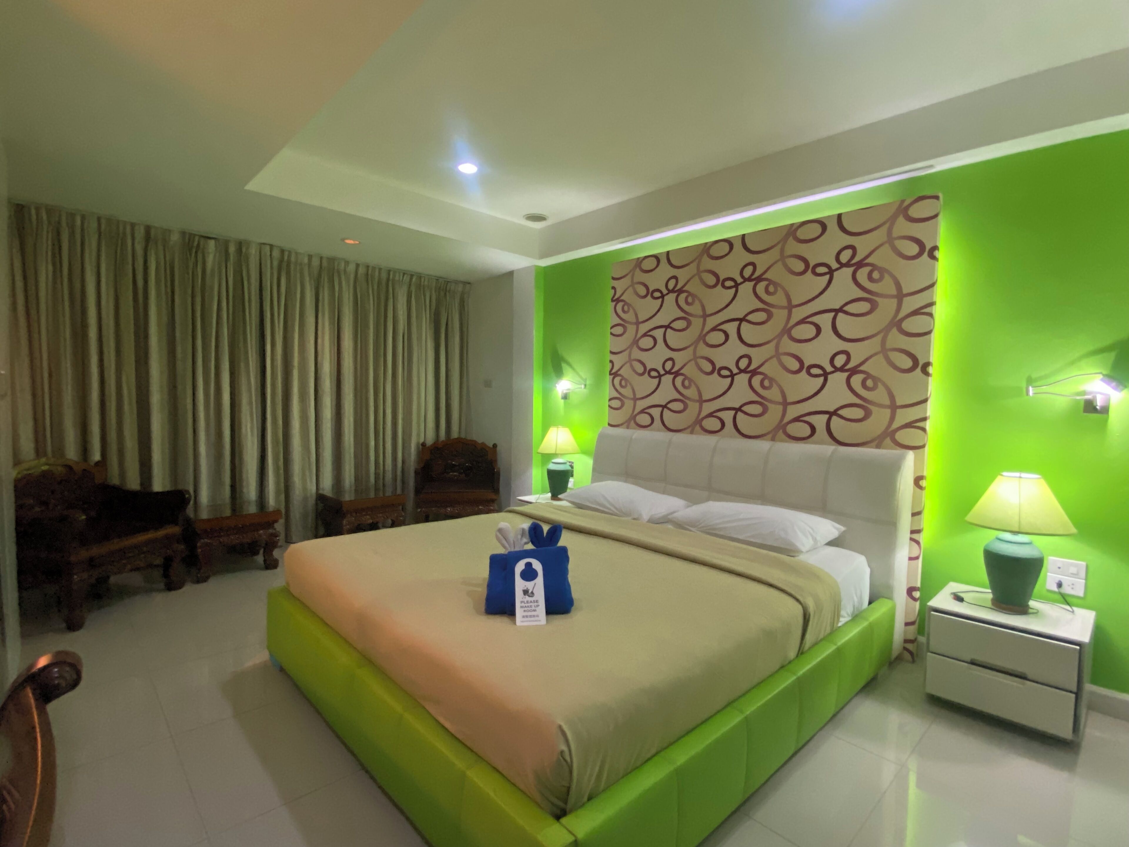 A hotel room with a bed, two nightstands, a green wall, a patterned wall, a wooden chair, and a curtain.