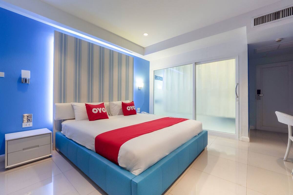 A modern hotel room with a large bed, a nightstand, a desk, and a sliding glass door leading to a balcony.