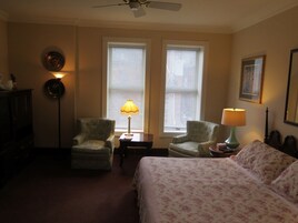 Habitación de hotel con dos sillones, cama con ropa de cama floral, ventilador de techo y dos ventanas con persianas.