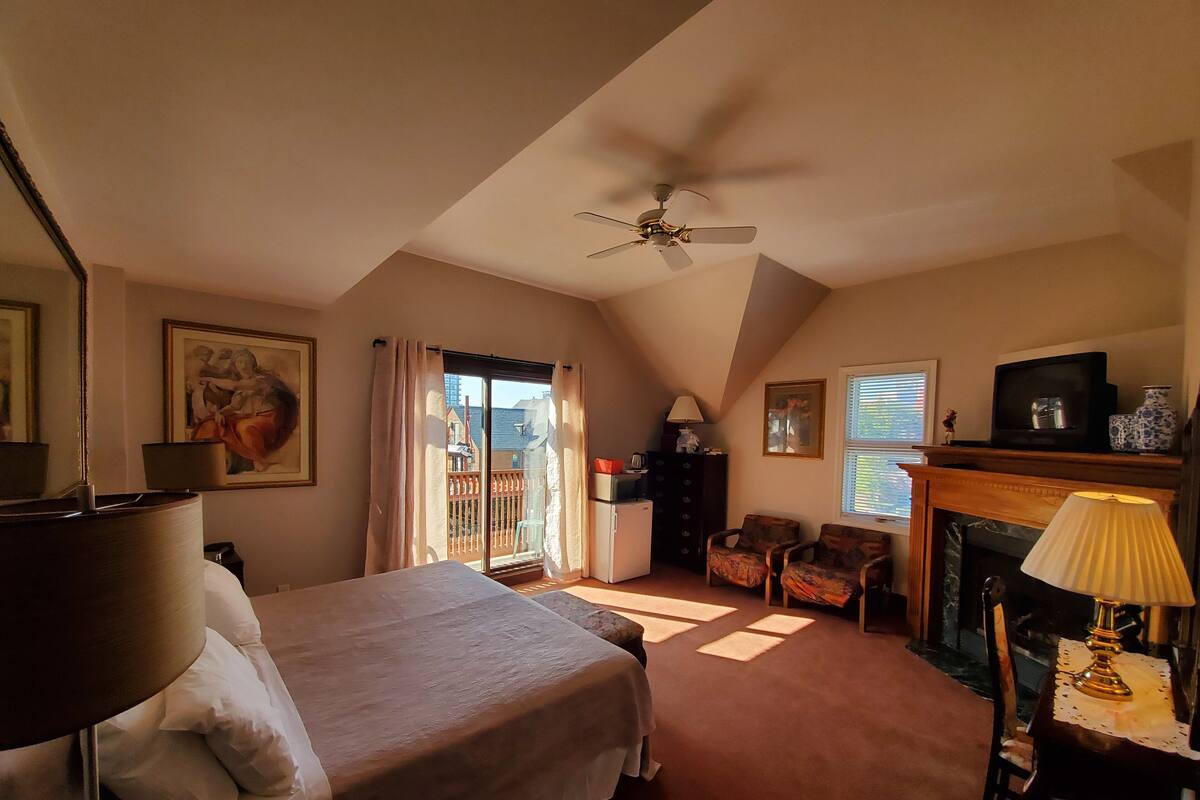 A bedroom with a bed, a ceiling fan, a fireplace, and a balcony with curtains.
