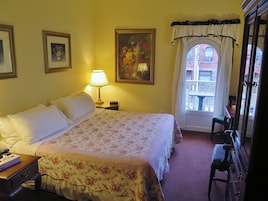 Een hotelkamer met een groot bed, een nachtkastje met een lamp, een lijst met bloemen, en een raam met uitzicht op een balkon.