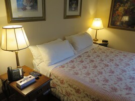 Habitación de hotel con cama, dos lámparas de noche, mesita de noche con teléfono y controles remotos, y una colcha con motivos florales.
