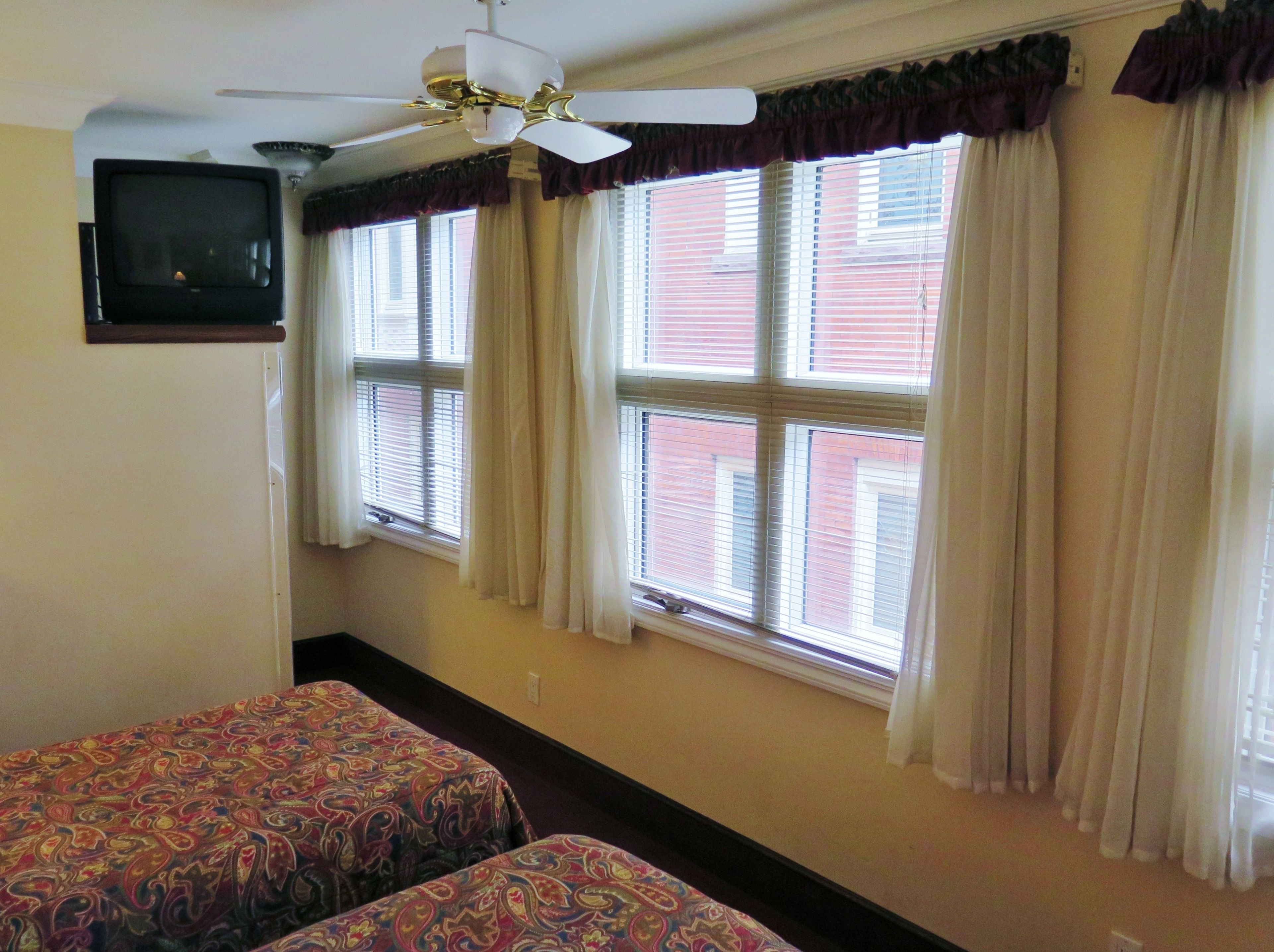 Una habitación con dos camas, ventilador de techo, televisión y ventanas con cortinas.