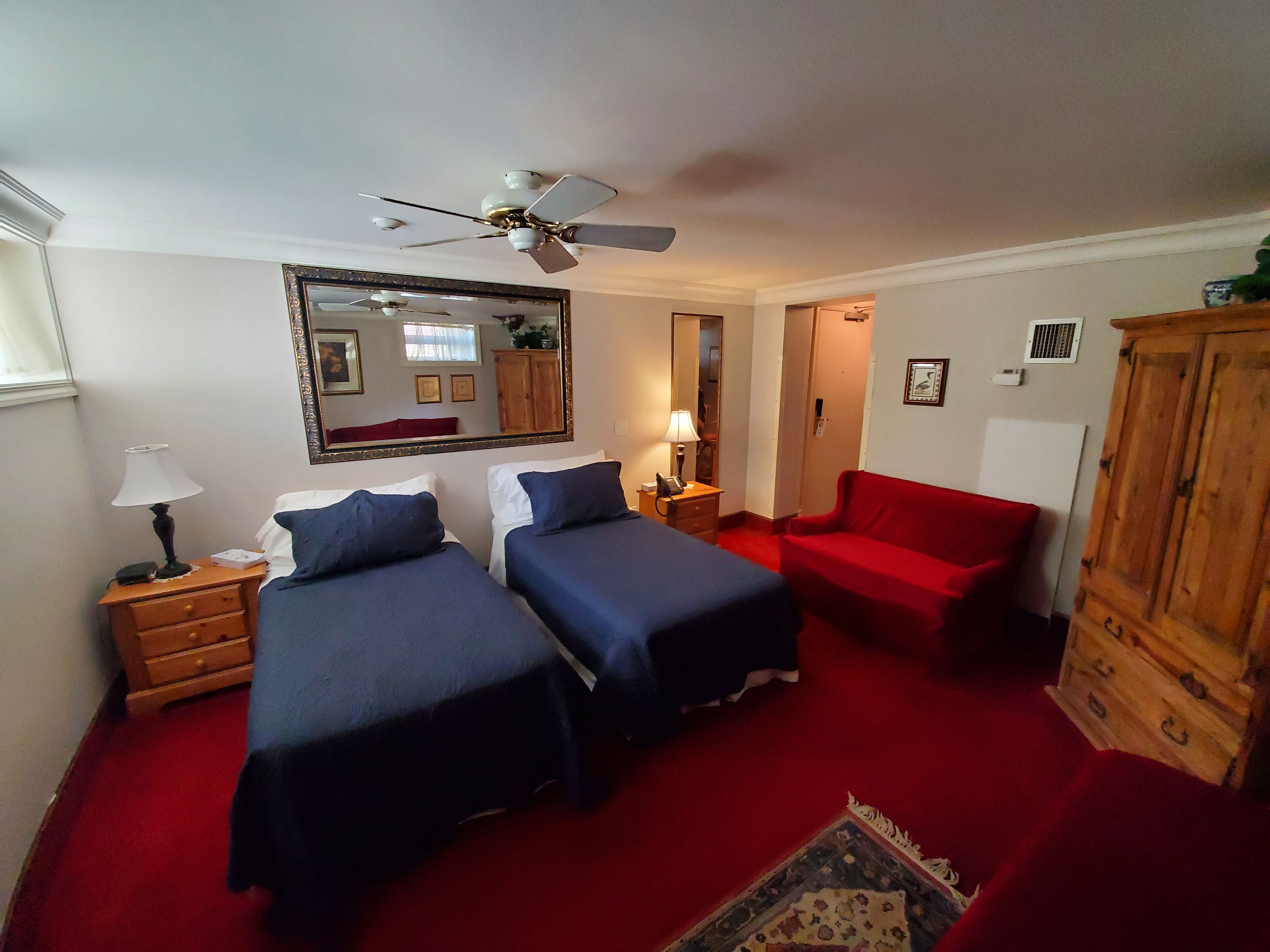 A room with two beds, a red sofa, a wooden cabinet, a ceiling fan, and a mirror on the wall.