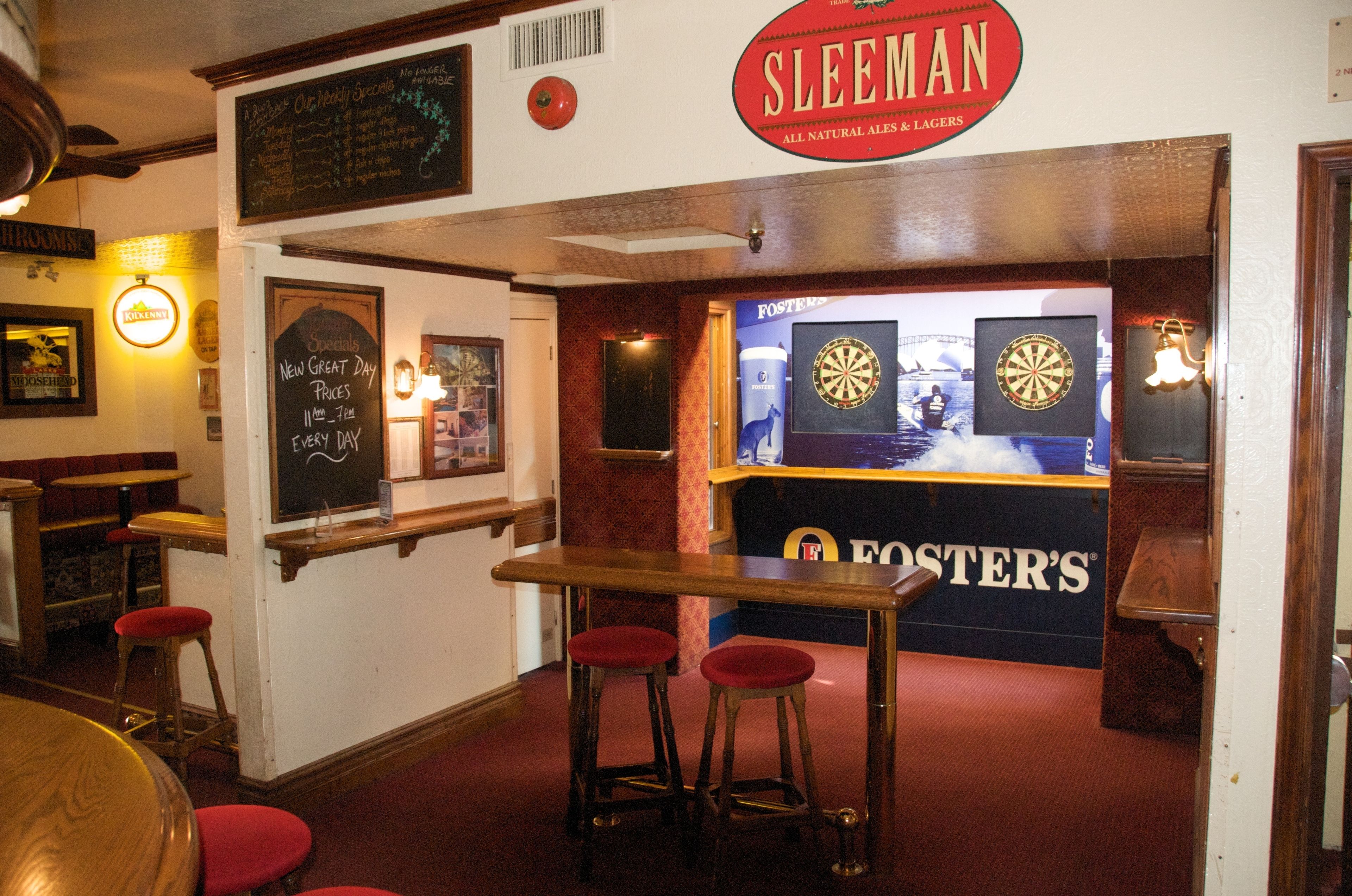 Zona de bar con taburetes, un dardo y un cartel de 'Foster's'.