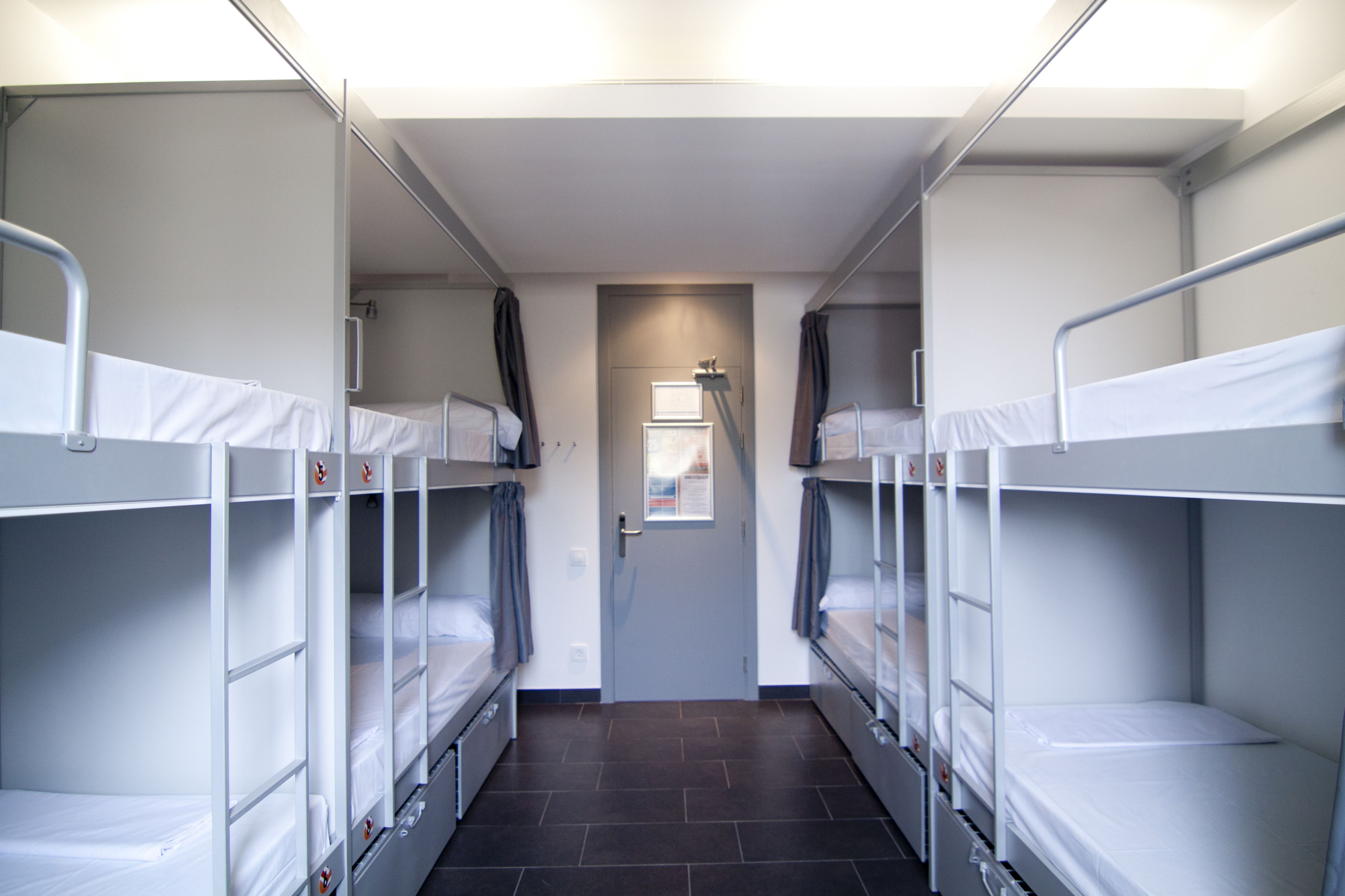 A narrow corridor with bunk beds on either side, a door at the end, and a window with curtains.
