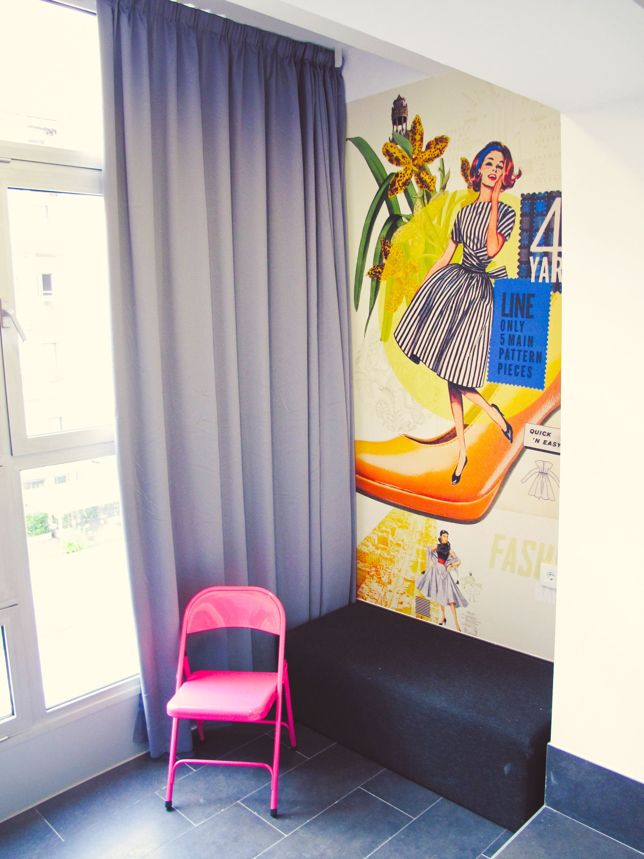 A room with a large window, a pink chair, a black sofa, and a wall mural.