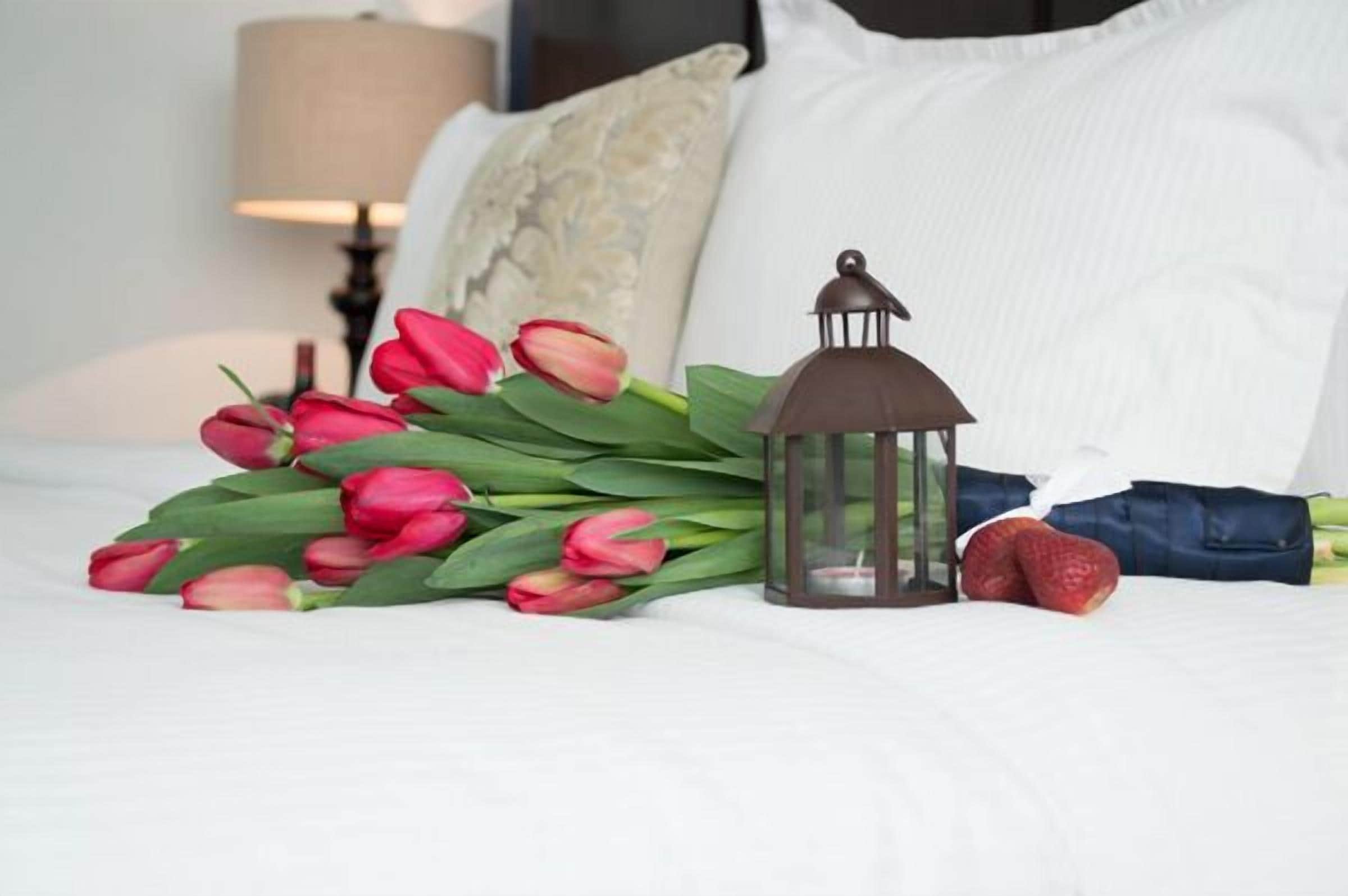 Ein Bett mit weißer Bettwäsche, ein Strauß roter Tulpen, ein Laternen und eine Erdbeere.
