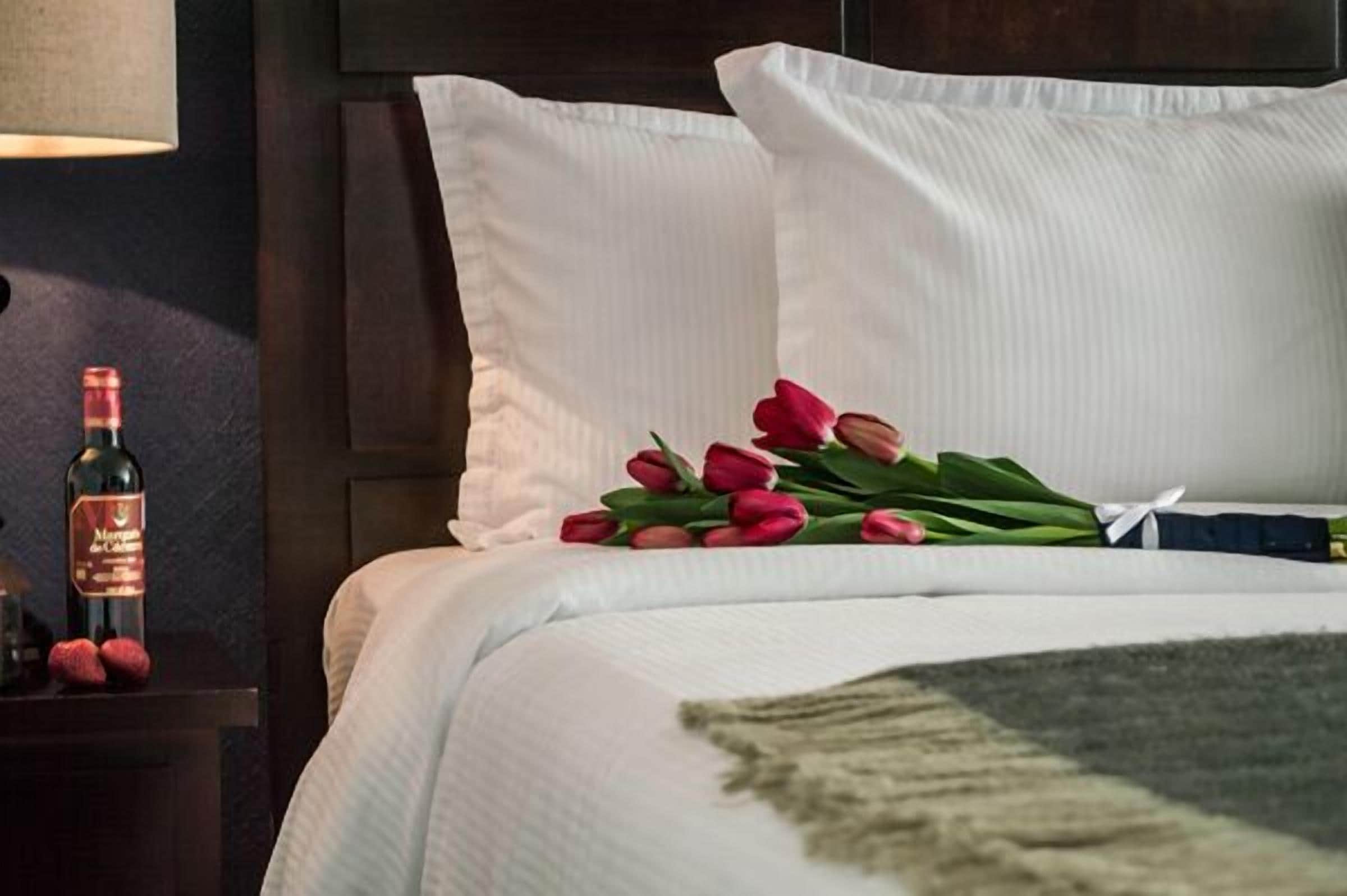 A neatly made bed with white linens, a bottle of wine, and a bouquet of red tulips on the nightstand.