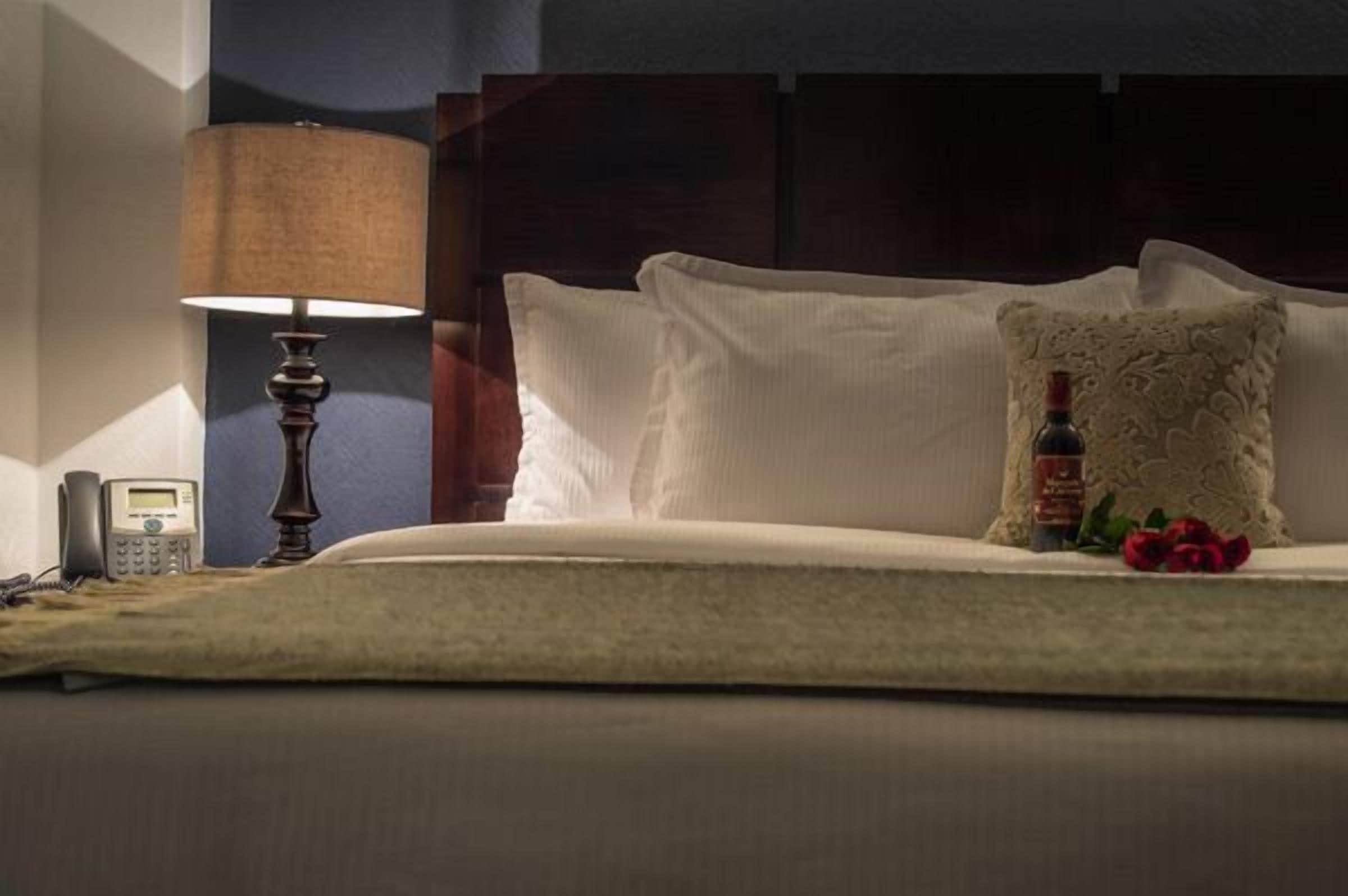 A neatly made bed with white linens, a lamp, a telephone, and a decorative pillow with a bottle and flowers.