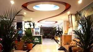 A hotel lobby with a 'WELCOME' mat, potted plants, a reception desk, and a TV screen.