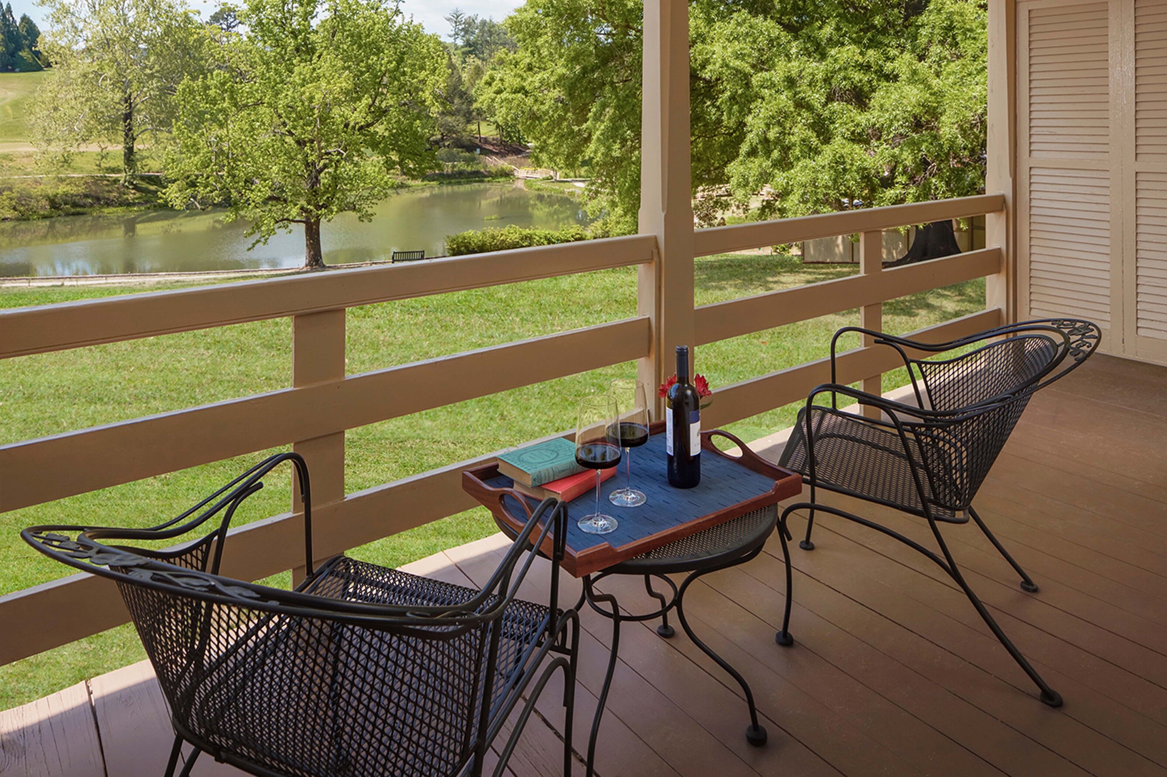 A covered outdoor seating area with two metal chairs and a table, overlooking a pond and greenery.