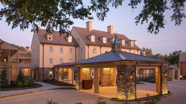 A well-lit gazebo with stone pillars and a tiled roof, surrounded by a landscaped area with a brick pathway leading to a building entrance. - Boar's Head Resort (Charlottesville)