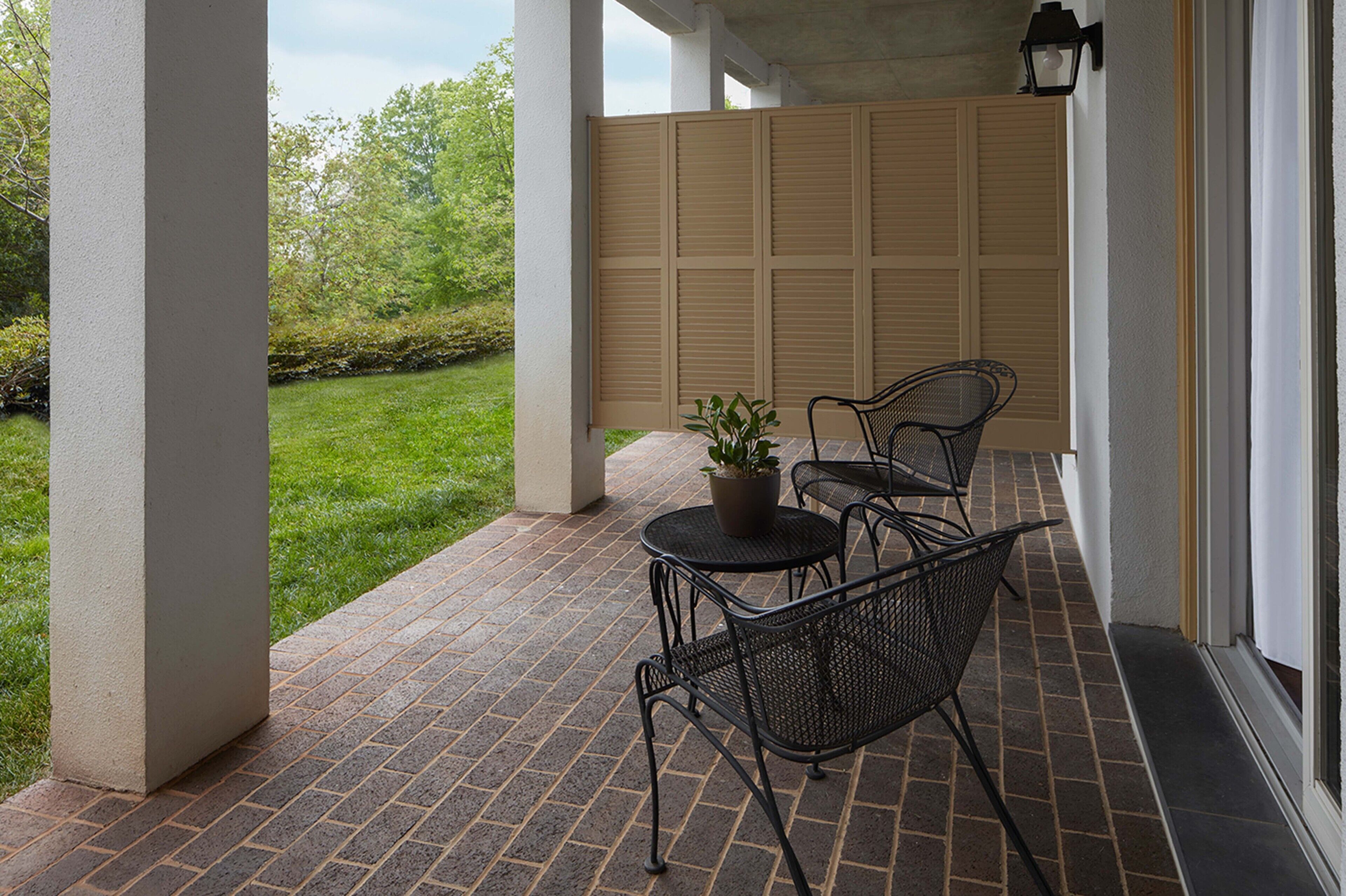 A covered patio with brick flooring, two metal chairs, a small round table, and a potted plant.