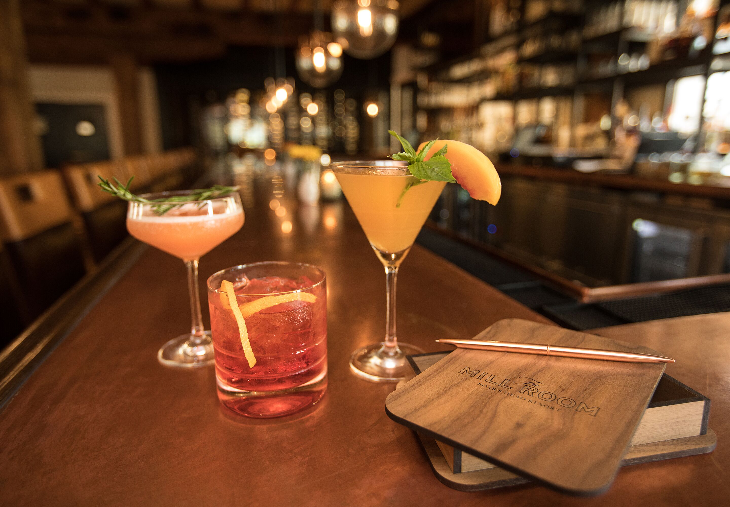 A bar area with two cocktails, a notebook, and a pen on the table.