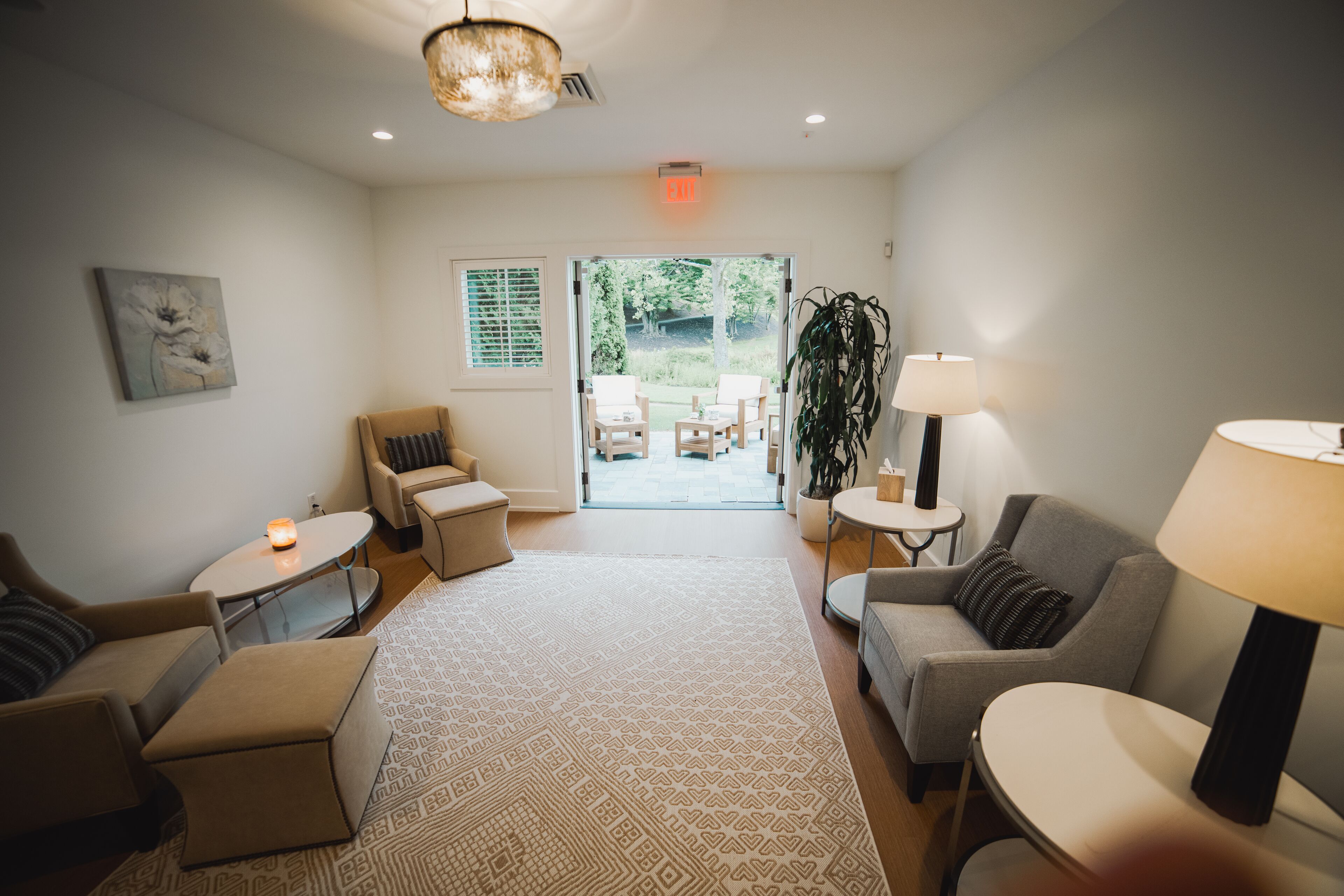 A modern living room with a sofa, armchairs, a coffee table, and a lamp. There is a door leading to an outdoor area with a table and chairs.