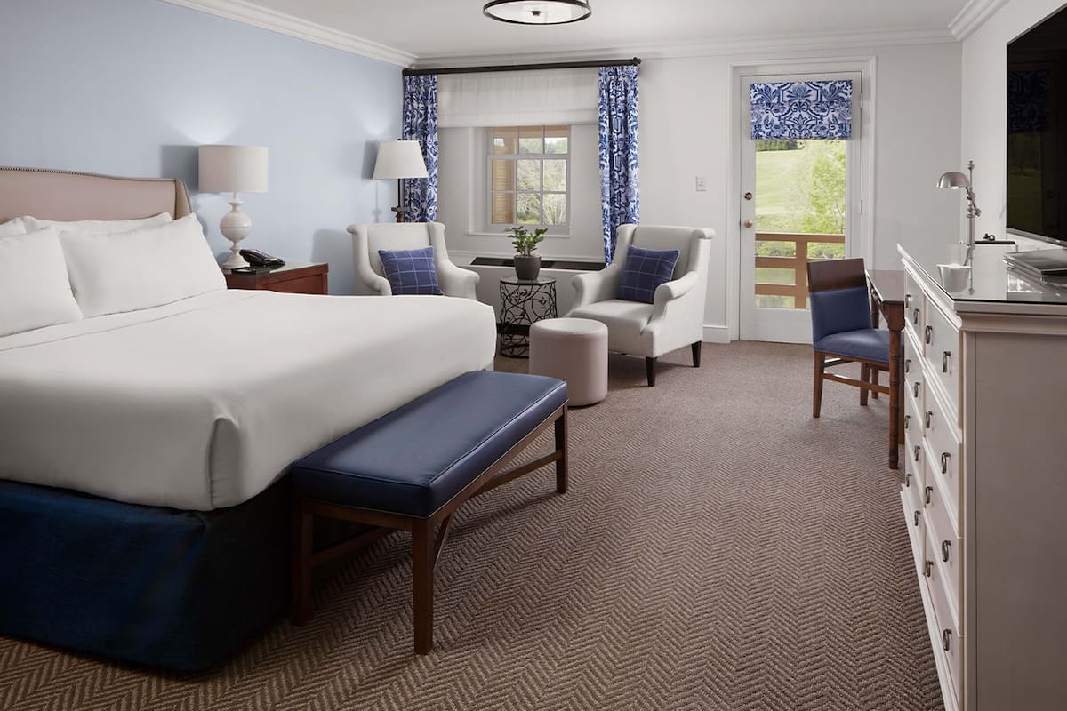 A hotel room with a large bed, two armchairs, a small table with a plant, a bench, and a dresser.