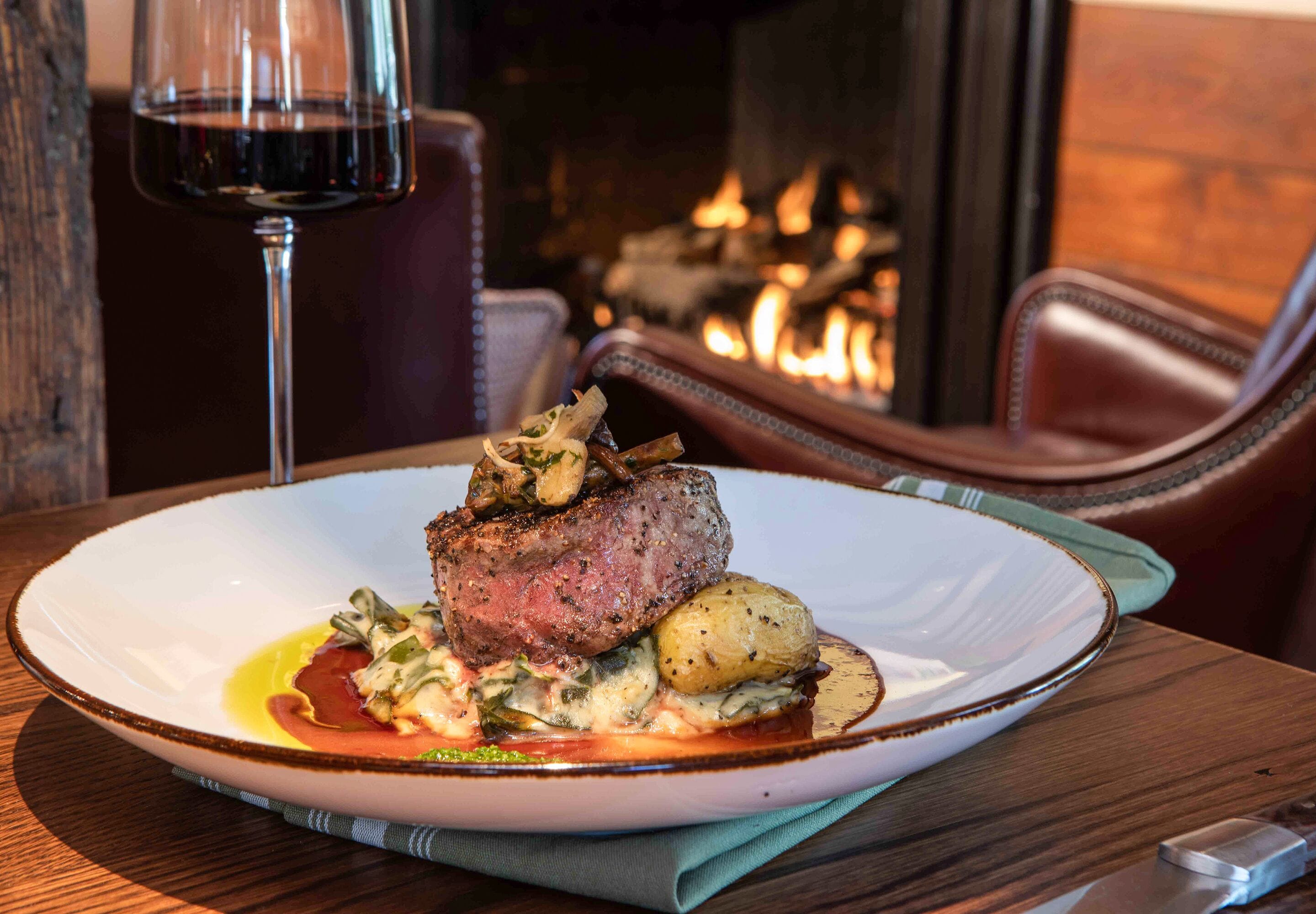 A plate of steak with vegetables and sauce on a table with a fireplace in the background.