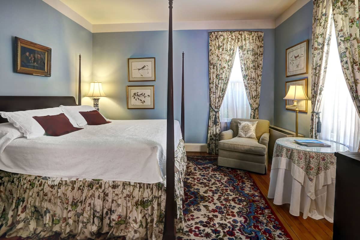 A bedroom with a four-poster bed, a chair, a small table, and a patterned rug.