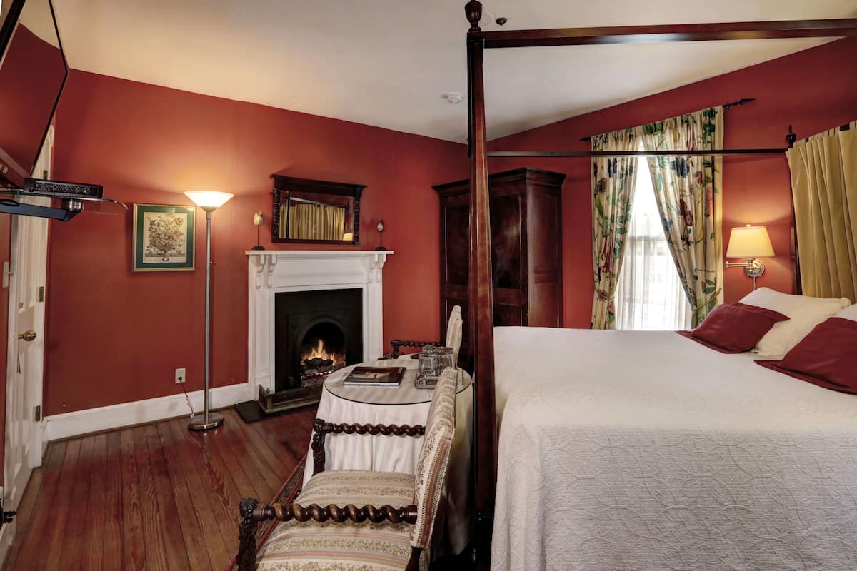 A room with a four-poster bed, a fireplace, a dining table with chairs, and a standing lamp.
