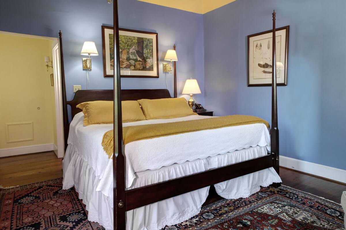 A four-poster bed with white bedding and a yellow throw, a patterned rug, and two wall-mounted lamps.