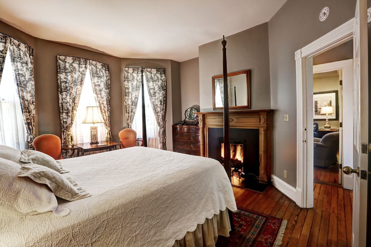 A bedroom with a bed, curtains, a fireplace, and a door leading to another room.