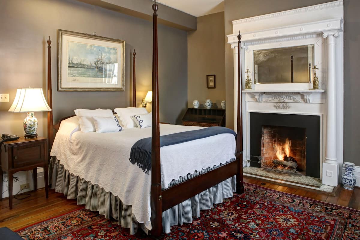 A four-poster bed with a white bedspread and a red and blue patterned rug. A fireplace with a white mantel and a mirror above it is visible.